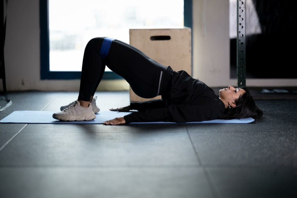 A woman in athletic wear lies on her back on a yoga mat, knees bent, feet flat on the floor, and lifts her hips in a bridge pose. She uses a resistance band above her knees and keeps her arms flat by her sides in a gym setting.