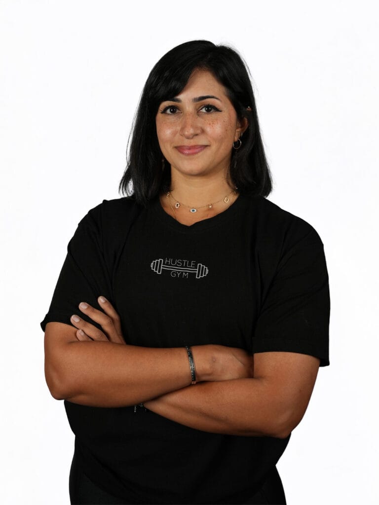 A woman with straight, shoulder-length black hair stands against a white background, smiling gently with her arms crossed. She wears a black T-shirt reading “Hustle Gym” with a barbell graphic, along with small hoop earrings and a choker necklace.