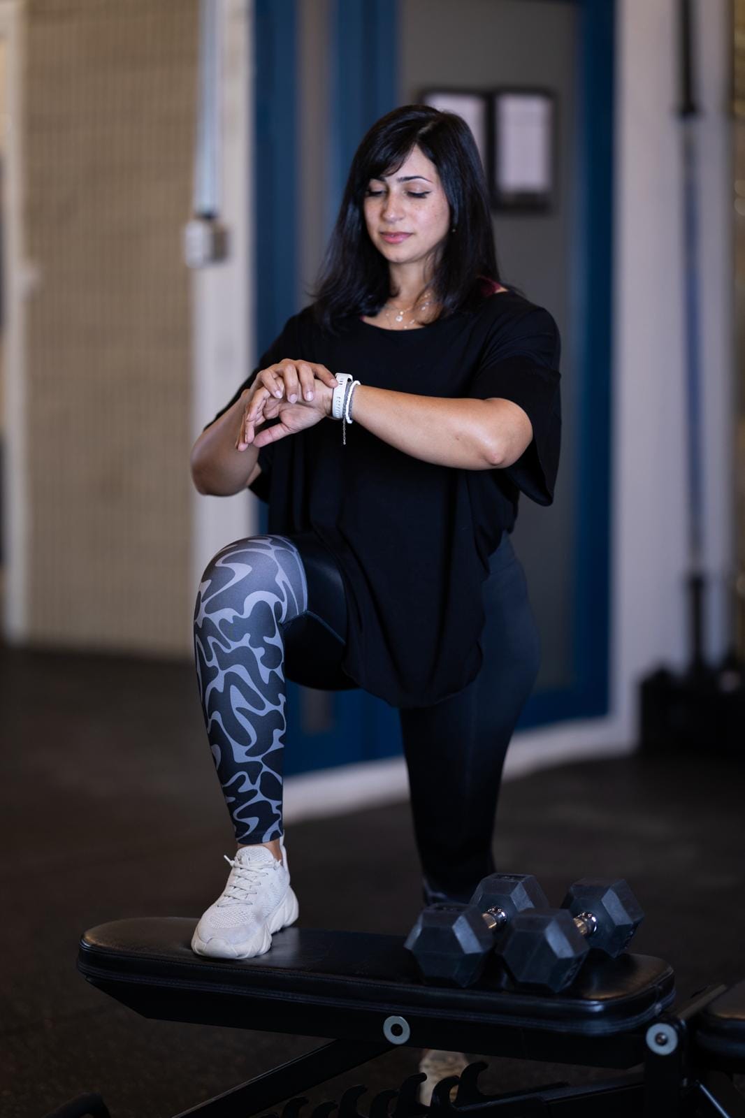 A woman in athletic wear kneels with one knee on a workout bench, checking her smartwatch. She wears a black shirt, patterned leggings, and white sneakers. Two dumbbells are on the bench in front of her. The gym background is out of focus.