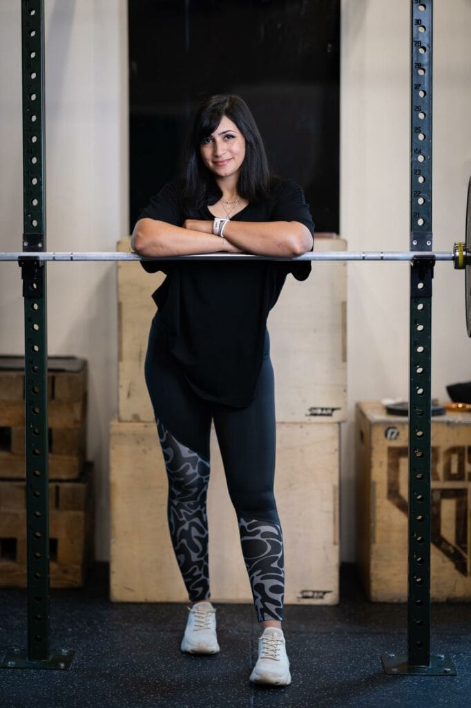 A woman with long dark hair leans her arms on a barbell resting on a squat rack in a gym. She wears a black shirt, patterned black leggings, and white sneakers. Wooden plyo boxes and weights are visible in the background.