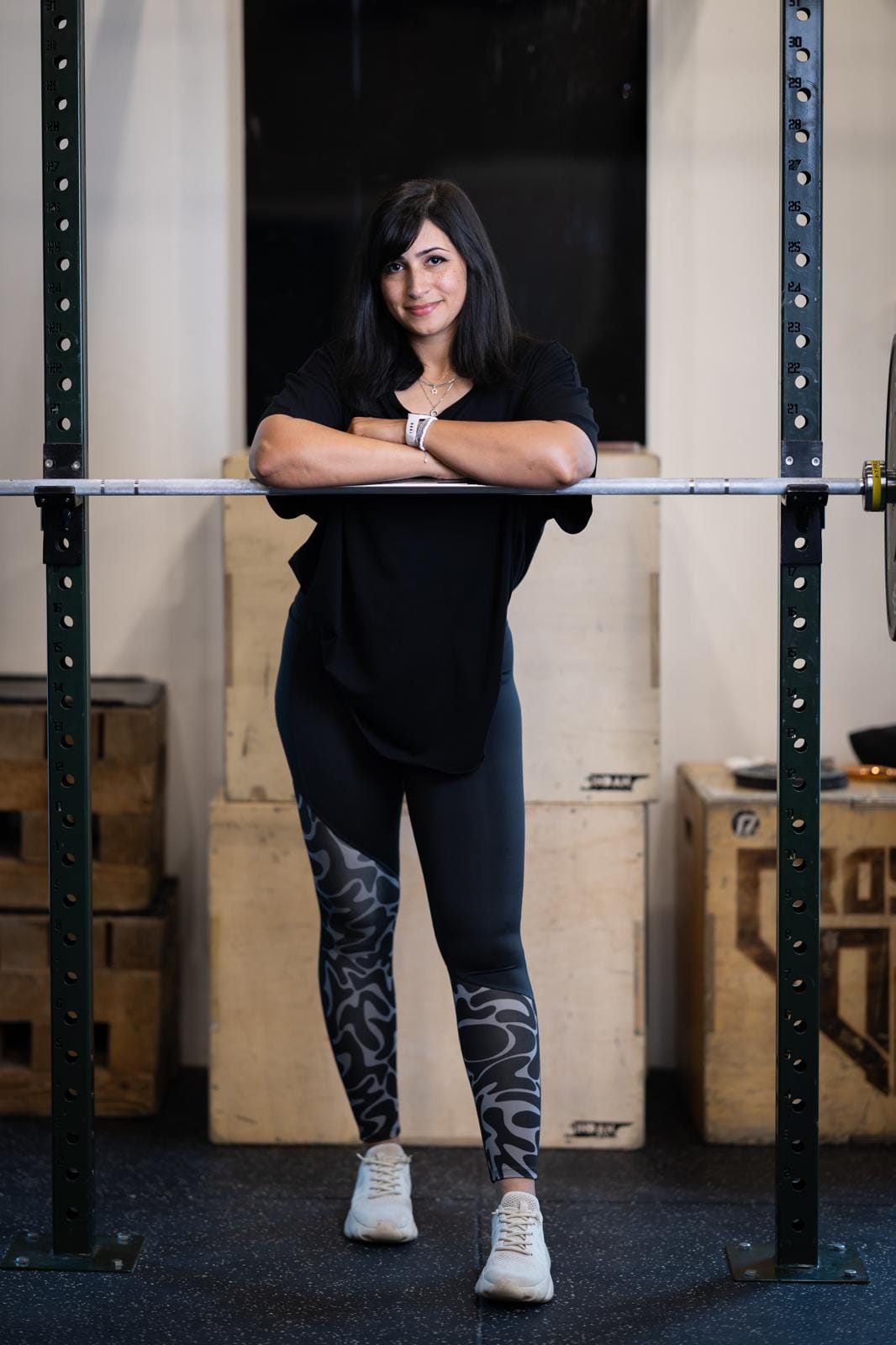 A woman with long dark hair leans her arms on a barbell resting on a squat rack in a gym. She wears a black shirt, patterned black leggings, and white sneakers. Wooden plyo boxes and weights are visible in the background.