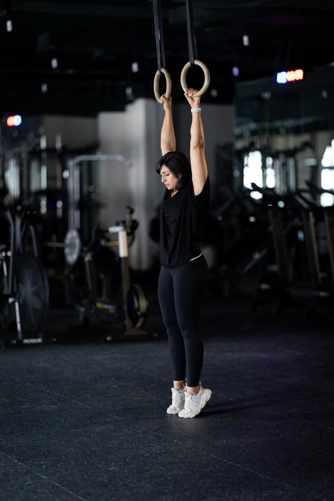 A woman in black athletic wear hangs straight-armed from wooden gymnastic rings in a dimly lit gym. She stands on tiptoe, keeping her feet together and body aligned, surrounded by gym equipment like bikes and weights in the background.