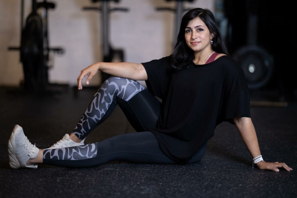 A woman with long dark hair sits on a gym floor, smiling at the camera. She wears a black loose shirt, patterned gray leggings, white sneakers, a bracelet, and small earrings. Gym equipment is blurred in the background.