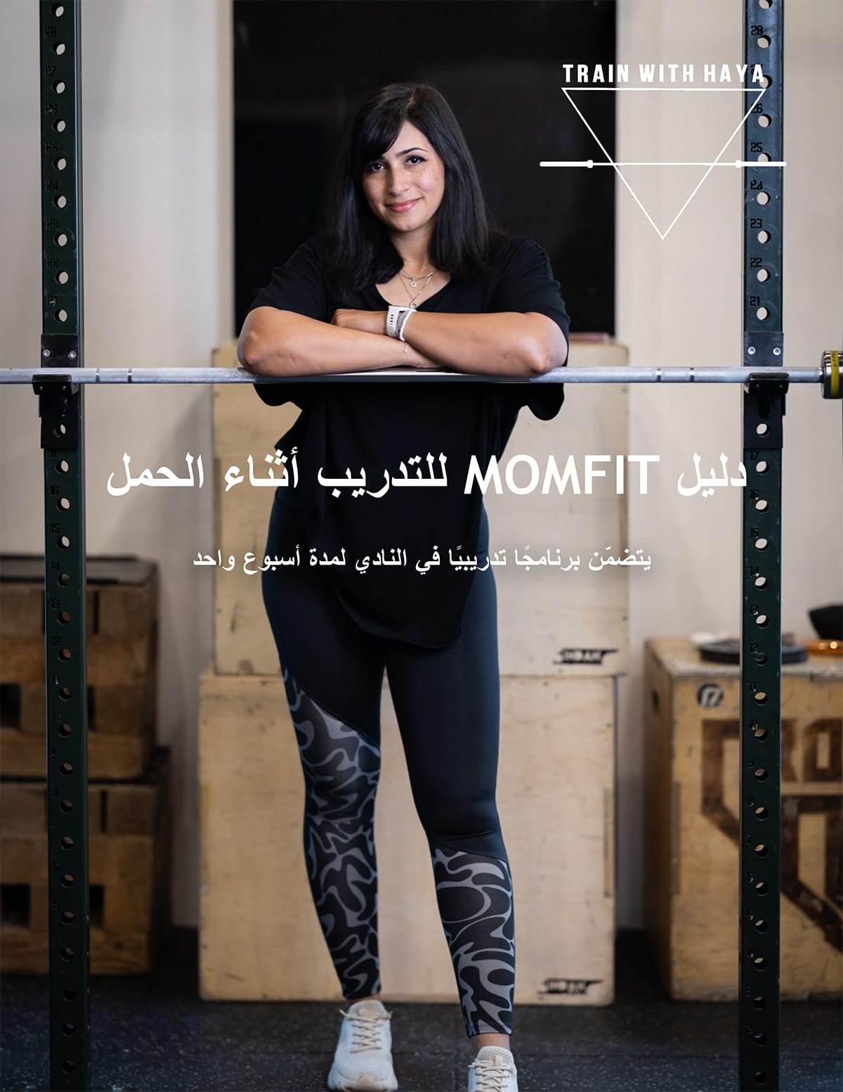 امرأة ذات شعر داكن طويل تقف بثقة في صالة الألعاب الرياضية، وتضع ذراعيها على حامل الأثقال. ترتدي قميصاً أسود وسروالاً ضيقاً منقوشاً. تظهر في الخلفية معدات الصالة الرياضية. يظهر على الصورة نص باللغة العربية وعبارة "تدربي مع حياة"، وهي تعلن عن دليل لياقة بدنية للحمل.